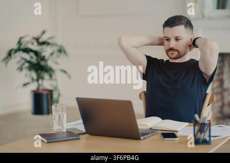 Caucasian man stretches with hands behind head while working on laptop in home office Stock Photo