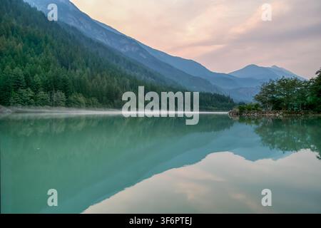 Mist drifts gently over tranquil lake surface. Village sits nestled on ...