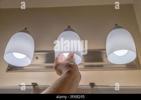hand replacing a light bulb in a modern bathroom vanity fixture with frosted glass shades. Home maintenance, electrical repair, DIY improvement and in Stock Photo