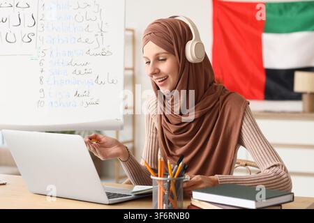 Smiling young muslim woman in hijab and headphones studying arabic language online with laptop at home, a whiteboard with arabic script and numbers is Stock Photo