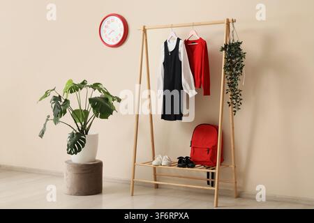 Childrens school uniform and red backpack on a wooden clothes rack with a monstera plant and red clock, a large monstera plant in a white pot sits on Stock Photo