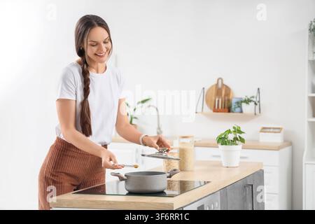 Smiling young woman cooking healthy food in a modern kitchen, adding spices to a pot on the stove Stock Photo