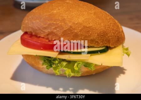 A sandwich sits on a white plate in a cafe. It has lettuce, tomato, cucumber, and cheese inside a round bun. The setting is casual and inviting. Stock Photo
