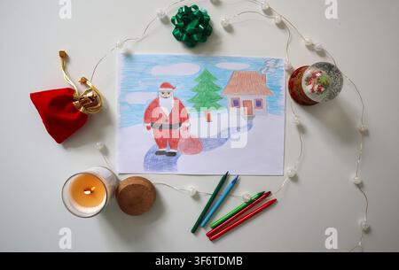 A child has drawn a picture of Santa Claus standing in front of a house with a tree. There are holiday decorations around the drawing, including a can Stock Photo