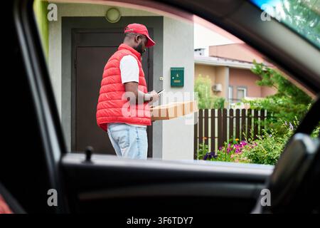 Black Courier Man Holding Cardboard Box and Using Smartphone near House Stock Photo