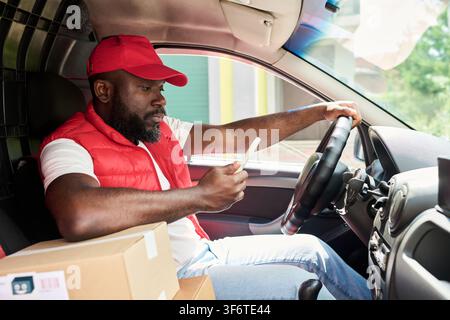 Black Courier Man Using Smartphone App While Driving Delivery Van Stock Photo