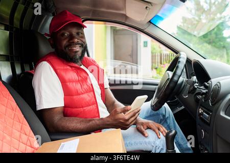Portrait of Black Man Working as Courier Holding Smartphone in Delivery Van Stock Photo