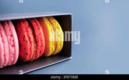 Close up colorful french macarons in row inside dark grey packaging box, with solid blue background. Sweet dessert pastry bakery food photography in m Stock Photo