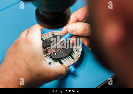 Technician repairs electronic circuit board using precision tools on a blue work surface with a microscope in the background Stock Photo