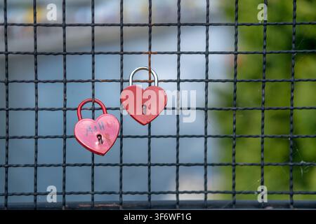 Two red heart padlocks are hanging on a black metal fence in front of a blurred background. Stock Photo