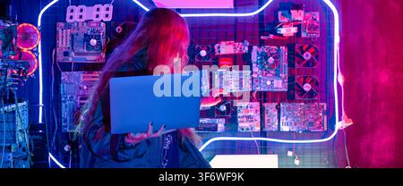 Asian woman in front of a computer monitor display big digital data with laptop computer, neon light, Hacker wear black jacket at system control room, Stock Photo