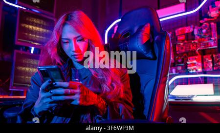 Asian woman in front of a computer monitor display using mobile phone, neon light, Hacker wear black jacket at system control room, Security guard wor Stock Photo