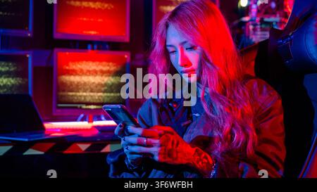 Asian woman in front of a computer monitor display using mobile phone, neon light, Hacker wear black jacket at system control room, Security guard wor Stock Photo