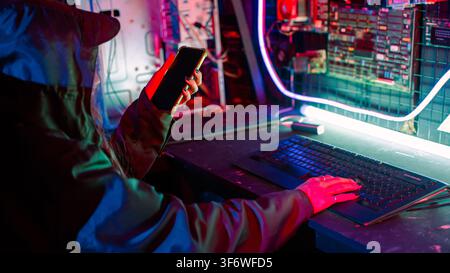 Woman hacker. Asian woman wearing hood working at computer and mobile phone typing coding hacking programming system. Hacker browsing internet uses ma Stock Photo