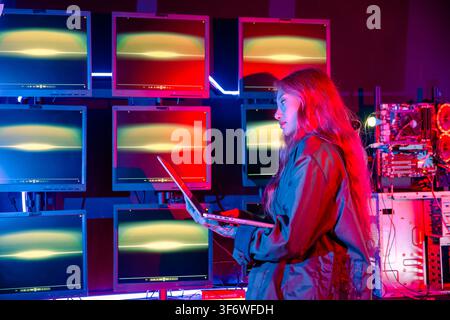 Asian woman holding laptop computer with CCTV room she check maintain security in monitor display, Hacker wearing a black jacket and has long red hair Stock Photo