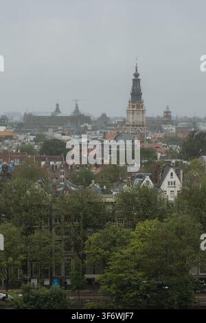 Amsterdam skyline capturing the Zuiderkerk bell tower rising above traditional Dutch canal houses and green trees under a cloudy sky Stock Photo