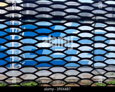 Abstract close-up of a black painted expanded metal mesh fence with diamond/hexagonal pattern. Blue and white modern building facade visible through t Stock Photo