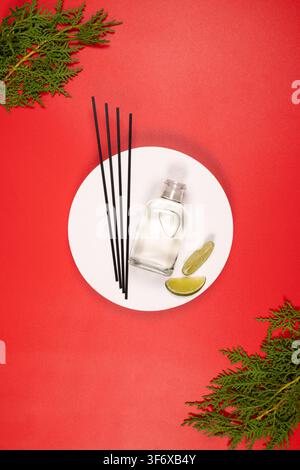 Reed diffuser with essential oil, black sticks, lime slices, and evergreen branches creating a fresh aroma and tranquil ambiance on a vibrant red back Stock Photo