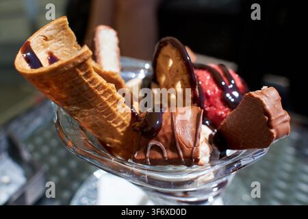 Close-up of a delicious ice cream sundae served in a glass bowl, topped with rich chocolate sauce, crispy wafer rolls, chocolate candy pieces, and fre Stock Photo