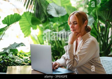 Pensive woman working on laptop surrounded by plants Stock Photo