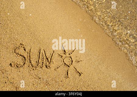Close up view of the word sun and a hand drawn sun symbol on wet golden sand at the seashore. Gentle ocean waves wash onto the beach in the background Stock Photo