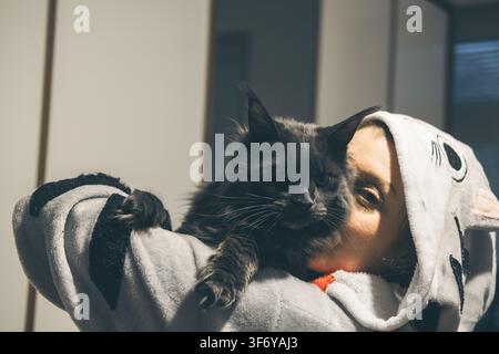 A young Caucasian woman with short hair holds a large black cat close to her face. She wears a cozy hoodie with animal patterns. The background is Stock Photo