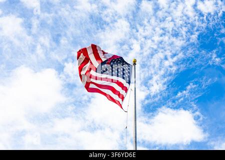 United States national flag waving on a metal flagpole against a bright blue sky with textured white clouds in broad daylight Stock Photo