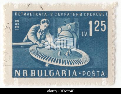 BULGARIA — 1961 June: 1.25 lev dull blue postage stamp depicts a skilled male machinist operating heavy industrial machinery inside a manufacturing plant. The worker is focused on precisely machining a large metal cogwheel using a specialized lathe or milling machine. This artwork forms part of a political propaganda series promoting the early fulfillment of the Third Five-Year Plan and celebrates socialist industrialization, heavy engineering, factory labor, mechanical production, and the rapid economic modernization goals in the People's Republic of Bulgaria Stock Photo