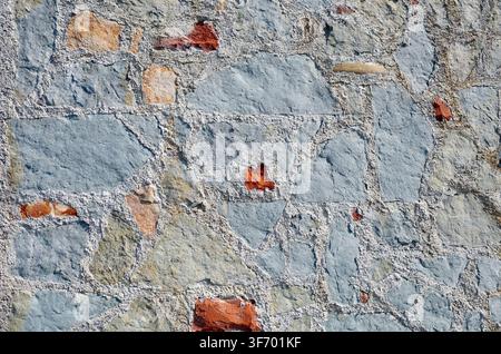 Detail of an old rustic natural stone wall and broken bricks in cement mortar. Cold blue grayish color with insert of ochre yellow and red. Background Stock Photo