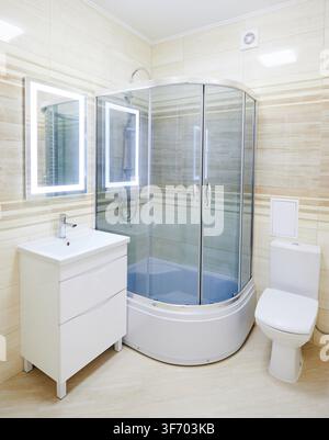 Modern bathroom interior with shower cabin, sink vanity and toilet. Bright washroom with illuminated mirror, beige tiles, glass doors and clean minimalist design Stock Photo