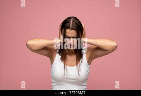 Young woman suffering from headache, covering ears with hands, feeling pain and frustration against a pink background Stock Photo