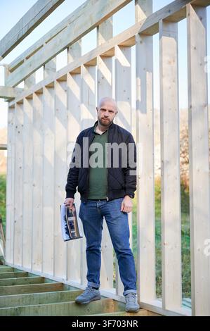 Builder stands inside timber wall frame holding project plans, overseeing construction progress and visualizing future layout of residential building Stock Photo