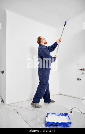 Builder applying white paint overhead using long handle roller. Clean modern interior under renovation, surface finishing and home improvement concept Stock Photo