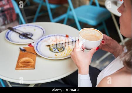 Woman holding coffee cup at cafe table after meal, empty plates and cutlery visible, close-up of hands with red nails, concept of finished breakfast, relaxation and casual dining Stock Photo