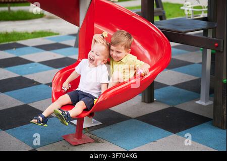 Two boys sliding together on red playground slide, laughing and holding toys, joyful outdoor play, park setting, happy childhood friendship moment Stock Photo