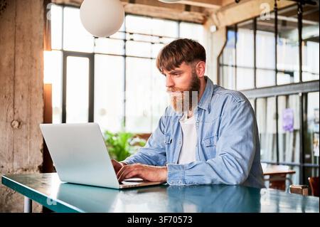 Bearded man working on laptop at high table in modern loft cafe, focused on screen in bright interior with glass walls and contemporary workspace atmosphere Stock Photo