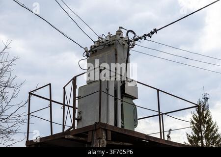 Metal power transformer unit stands on an elevated outdoor structure connected to overhead power lines, illustrating energy supply, electrical enginee Stock Photo