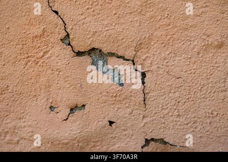 Close-up background texture of damaged peach orange wall with cracked plaster and peeling paint showing underlying concrete. Stock Photo