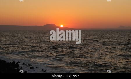 Sun setting directly behind the coastal cape and headland seen from Rethymno beach, Crete, Greece. Stock Photo