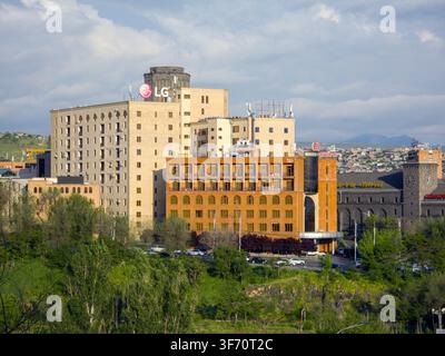 Large urban building complex on hillside. Green trees surround base of structure. Sky has scattered clouds. Distant mountains visible in background Stock Photo