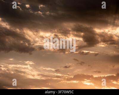 Dramatic sunset sky with golden clouds and dark storm clouds. Warm light breaks through cloud layers. Textured clouds create moody atmosphere Stock Photo