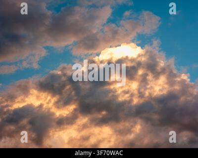 Sunlight breaks through dramatic clouds at sunrise or sunset. Warm golden light highlights cloud edges against deep blue sky Stock Photo