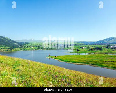 Sunlit lake nestled in green valley between rolling hills. Clear blue sky above calm water surface. Grasses and wildflowers grow along shoreline Stock Photo