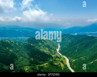 Winding road cuts through lush green mountains under bright blue sky. Scenic view with rolling hills covered in dense vegetation Stock Photo