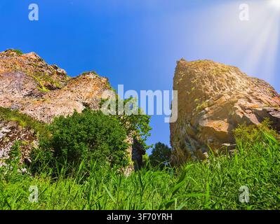 Two rocky cliffs rise under bright blue sky. Green grass and bushes grow at base. Sunlight shines from behind right cliff Stock Photo