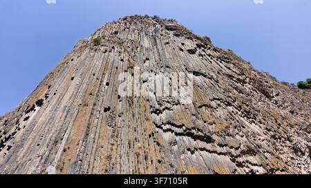 Steep, layered cliff face rises sharply against clear blue sky. Rock surface shows vertical striations and weathered texture Stock Photo