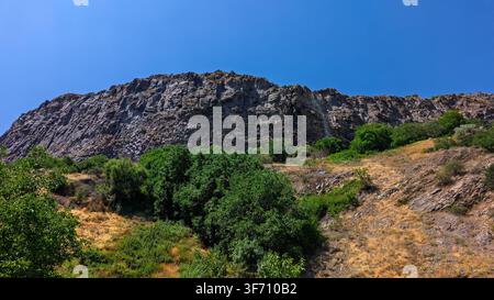 Rugged rocky hillside under clear blue sky. Green shrubs grow on dry, sunlit slope Stock Photo