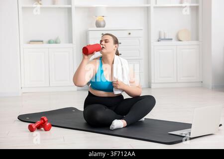 Overweight woman drinking water on fitness mat at home Stock Photo