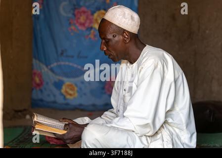Muslim man in white robe and cap reads Quran quietly. He sits cross-legged on green mat, focused and peaceful. Blue floral cloth hangs behind him. Sce Stock Photo