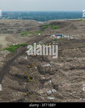 Aerial view of a large landfill site with multiple yellow excavators working on piles of waste, surrounded by sparse vegetation and a hazy horizon in Stock Photo